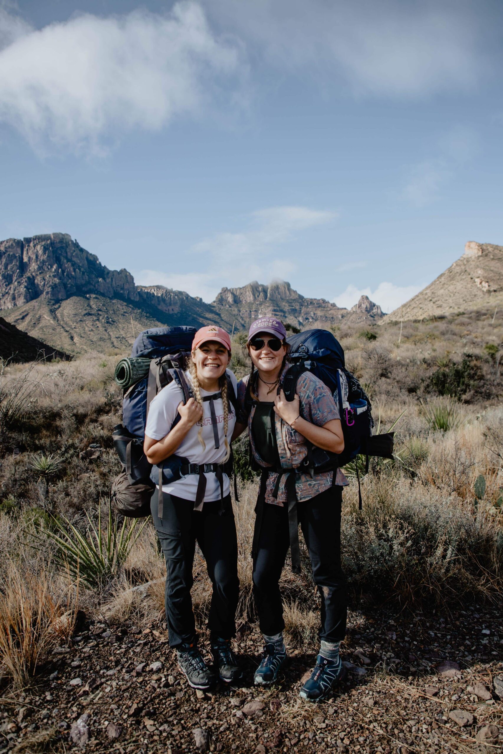This is a picture of two girls with a lot of hiking gear on. They are in front of mountain with big backpacks on.