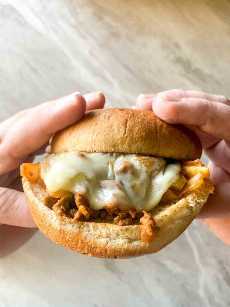 These is a hand holding up a sloppy joe. There is a sloppy joe with a toasted bun, mix, and cheese.