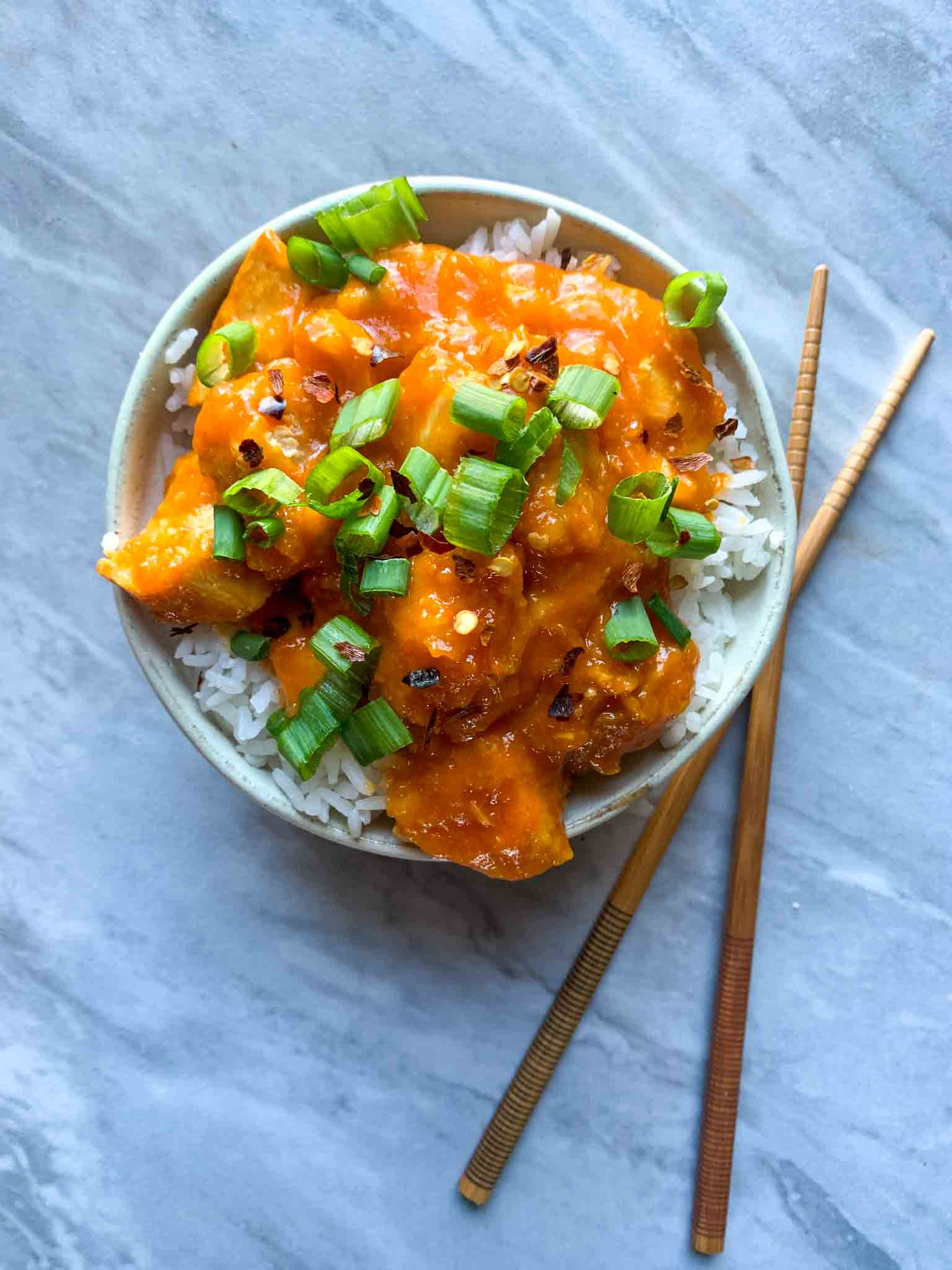 This is a bowl of firecracker chicken. There is a cream colored bowl with white rice and chunks of bright orange chicken on top. On the chicken is cut green onions and red pepper flakes. A pair of chopsticks is to the right of the bowl.