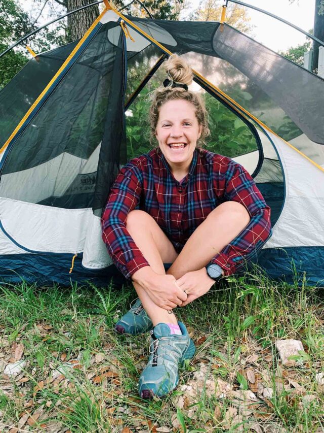This is a girl in a red and blue flannel shirt sitting in the opening of a tent. She is smiling with her arms wrapped around her legs.