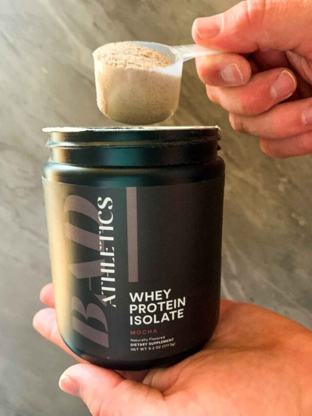 This is a picture of a jar of protein powder. This is a hand holding a scoop of the protein powder.