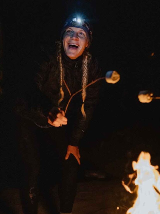 This is a picture of a girl wearing all black with a headlamp on. She is roasting a marshmallow on the campfire.