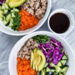 This is a picture of spring roll bowls. There are two white bowls filled with the spring roll combinations. Each bowl has a section of white rice, shredded carrots, turkey, purple cabbage, green cucumber, green avocado, green onions, and a sauce.