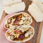 This is a bacon breakfast burrito. There is a tortilla being held up by a hand and it has eggs, cheese, and slices of bacon. There are a few wrapped burritos on a cutting board behind the hand.