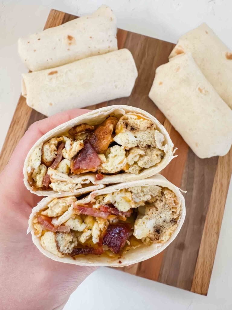 This is a bacon breakfast burrito. There is a tortilla being held up by a hand and it has eggs, cheese, and slices of bacon. There are a few wrapped burritos on a cutting board behind the hand.