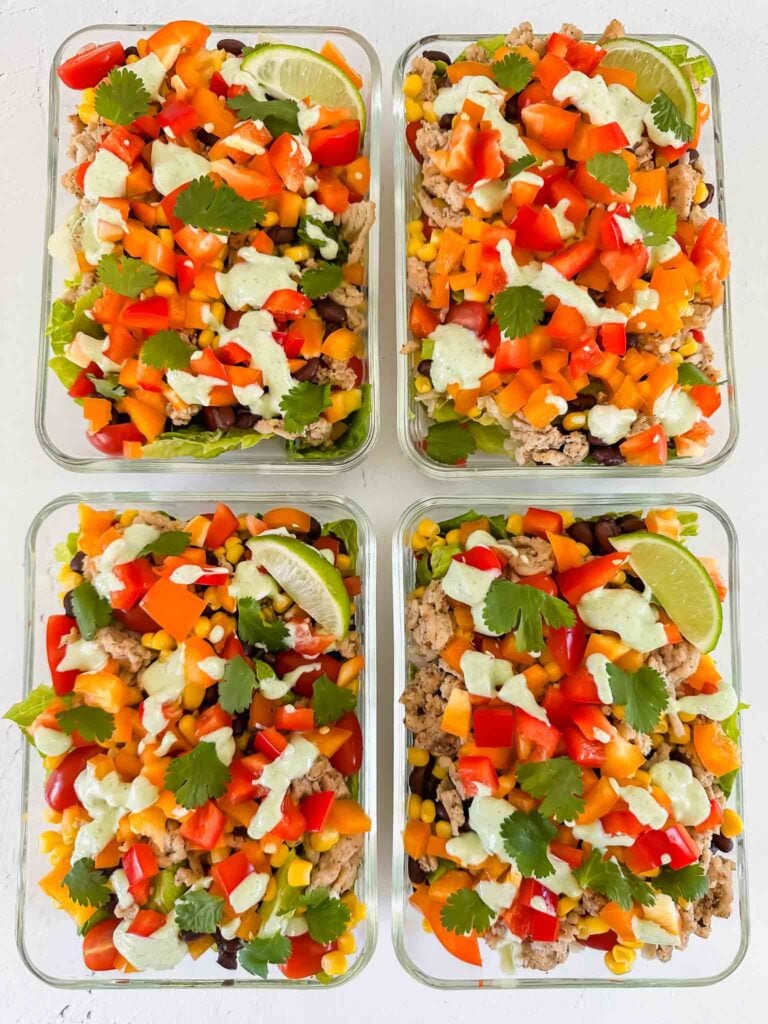This is a picture of turkey taco bowls. There are 4 glass meal prep containers filled with the bowls. There is cooked turkey, diced orange bell peppers, red tomatoes, corn, and black beans. The bowls are topped with chopped cilantro, a lime wedge, and a light green cilantro lime sauce.