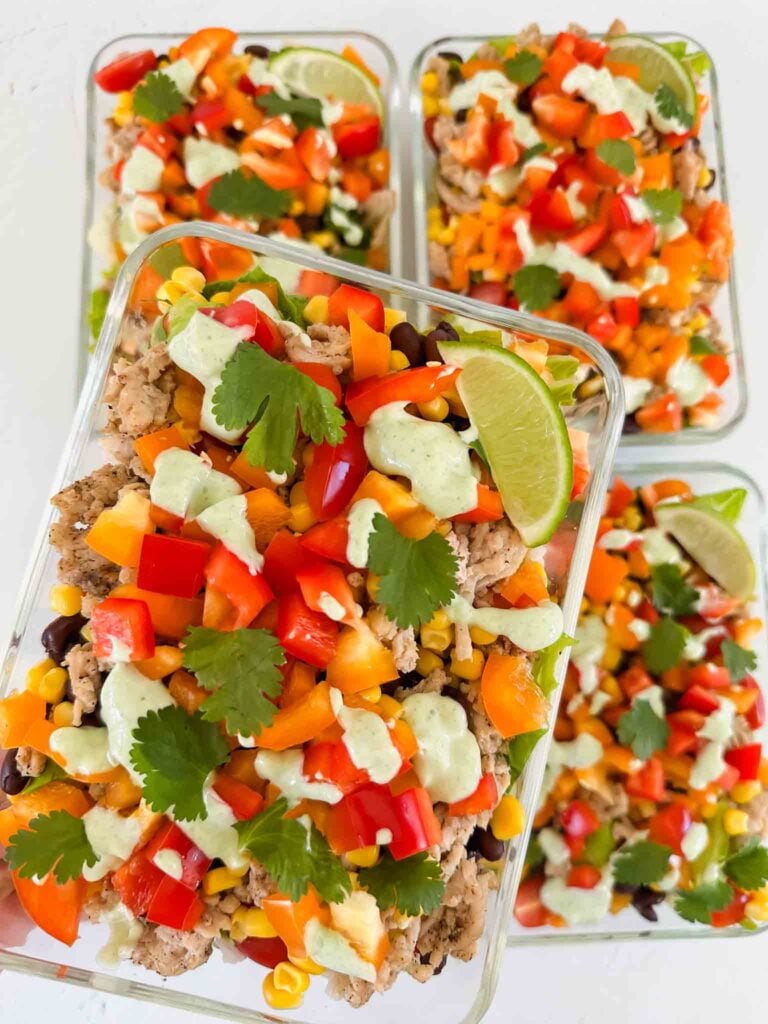 This is a picture of turkey taco bowls. There are 4 glass meal prep containers filled with the bowls. There is cooked turkey, diced orange bell peppers, red tomatoes, corn, and black beans. The bowls are topped with chopped cilantro, a lime wedge, and a light green cilantro lime sauce. A hand is holding up one of the meal prep containers.