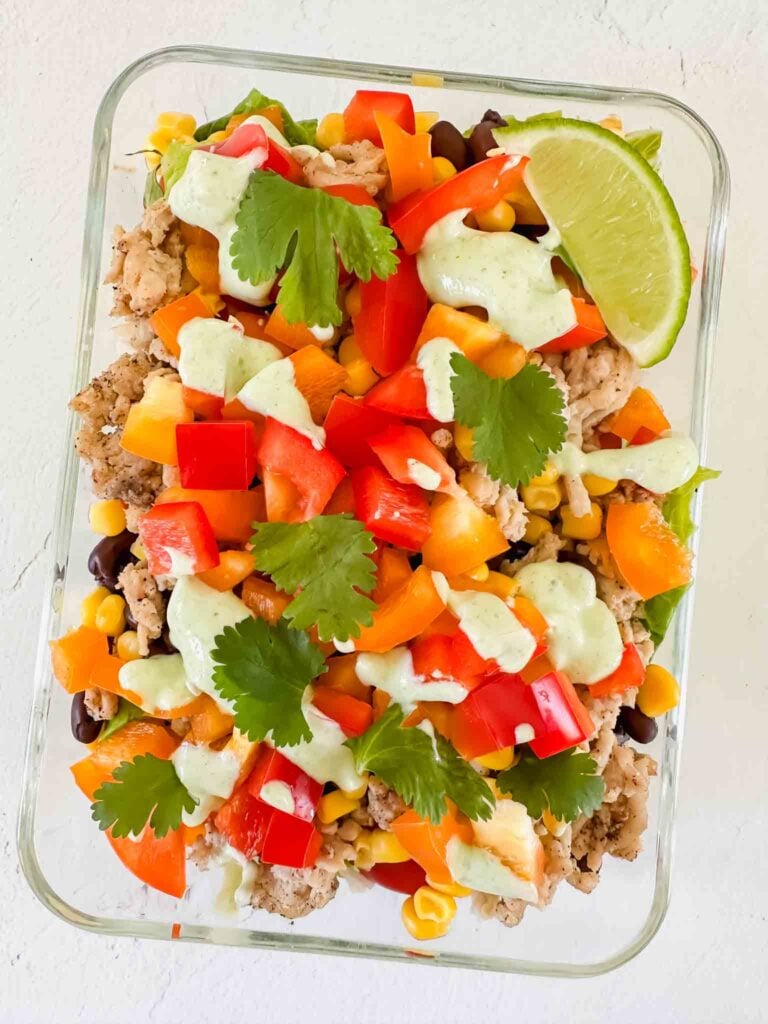 This is a picture of turkey taco bowls. There is a glass meal prep container filled with the prepped bowl. There is cooked turkey, diced orange bell peppers, red tomatoes, corn, and black beans. The bowls are topped with chopped cilantro, a lime wedge, and a light green cilantro lime sauce.