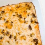 This is a picture of the breakfast hashbrown casserole. There is a white casserole dish filled with the baked item. There's a golden egg finish with pieces of brown sausage and it's topped with a dark yellow/orange melted cheese.