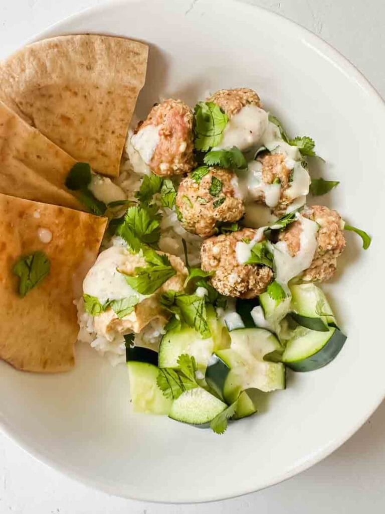 This is a picture of greek turkey meatballs. There's a bowl filled with white rice, greek meatballs, cucumbers, hummus, and pita. It's topped with a tzaziki sauce.