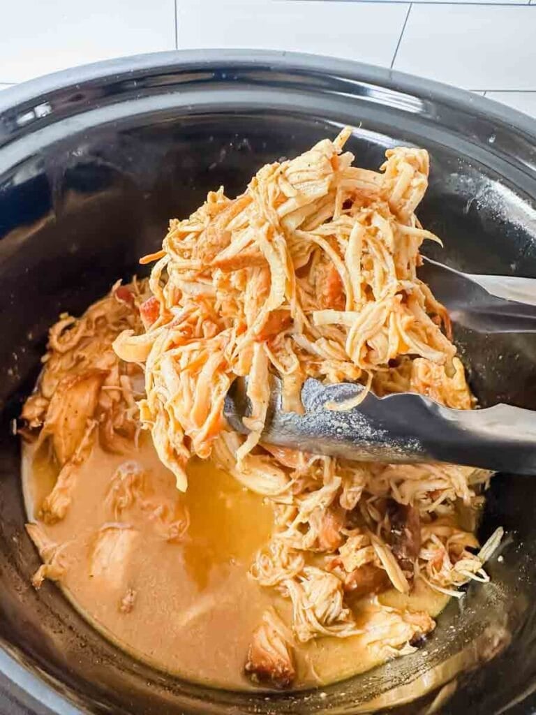 This is a picture of crockpot honey mustard chicken. There is large black crockpot filled with shredded chicken in a orange honey mustard sauce. There's silver tongs holding shredded chicken.