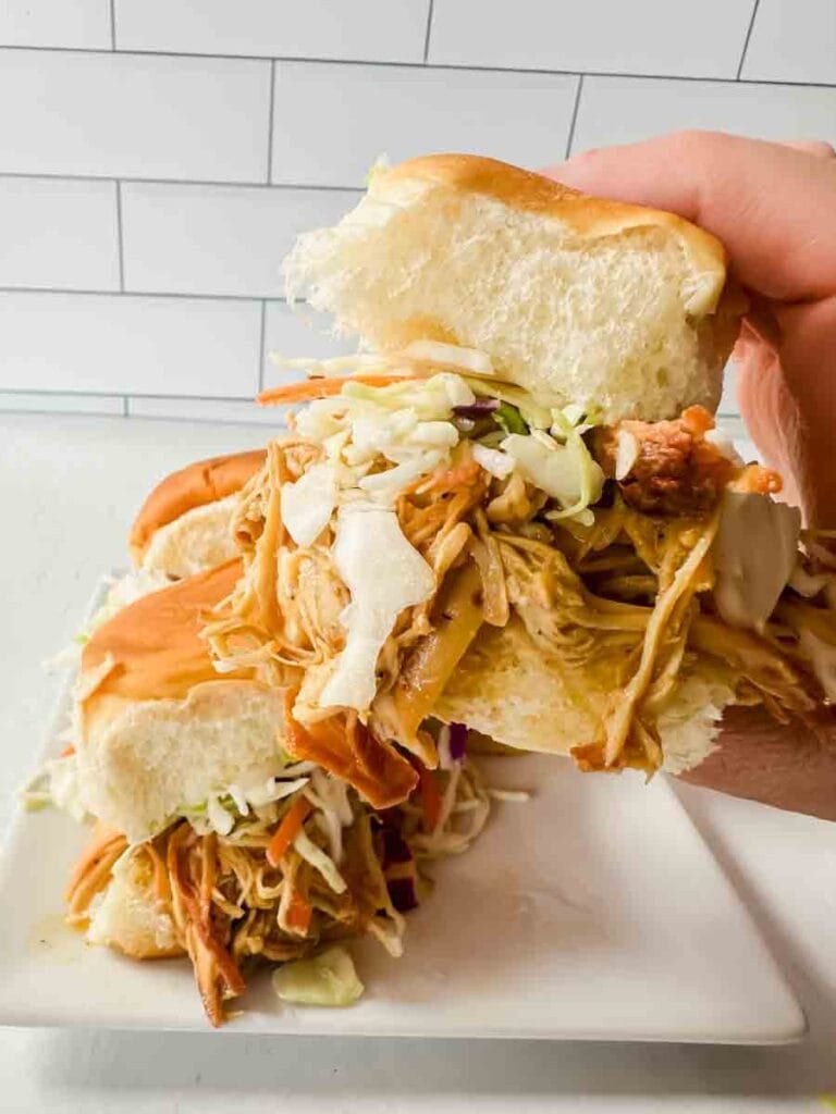 This is a picture of honey mustard sliders. There's a white plate holding sliders that have shredded chicken and slaw on buns. A hand is holding up a honey mustard chicken slider.