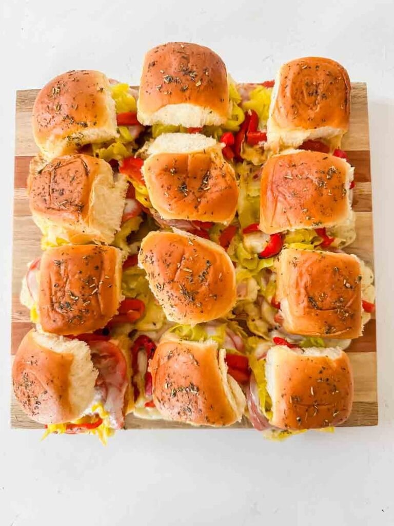 This is a picture of hot italian sub sliders. There's a cutting board with sliders stacked on them. On the sliders you can see melted cheese, salami, pepperoni, red roasted peppers, and banana peppers on top. The rolls are light brown and speckled with butter and italian season. They look perfect.