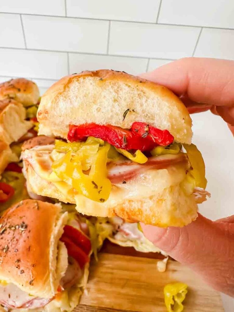 This is a picture of hot italian sub sliders. There's a hand holding up a hot italian sub slider. On the sliders you can see melted cheese, salami, pepperoni, red roasted peppers, and banana peppers on top. The rolls are light brown and look perfect.