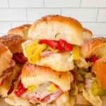 This is a picture of hot italian sub sliders. There's a cutting board with sliders stacked on them. On the sliders you can see melted cheese, salami, pepperoni, red roasted peppers, and banana peppers on top. The rolls are light brown and look perfect.