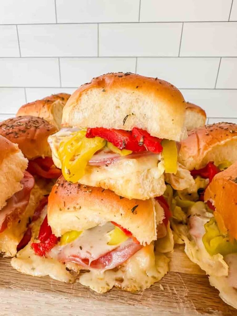 This is a picture of hot italian sub sliders. There's a cutting board with sliders stacked on them. On the sliders you can see melted cheese, salami, pepperoni, red roasted peppers, and banana peppers on top. The rolls are light brown and look perfect.