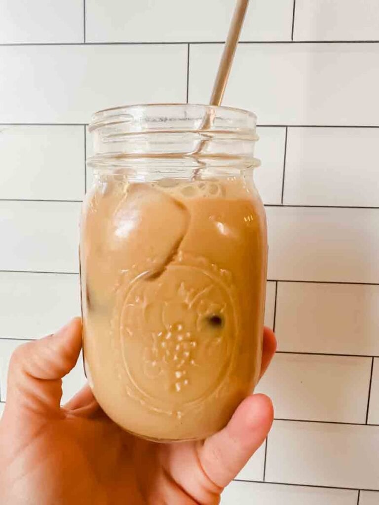This is a picture of an iced sf latte. There's a glass mason jar filled with a light brown latte and straw in the cup. A hand is holding up the glass.