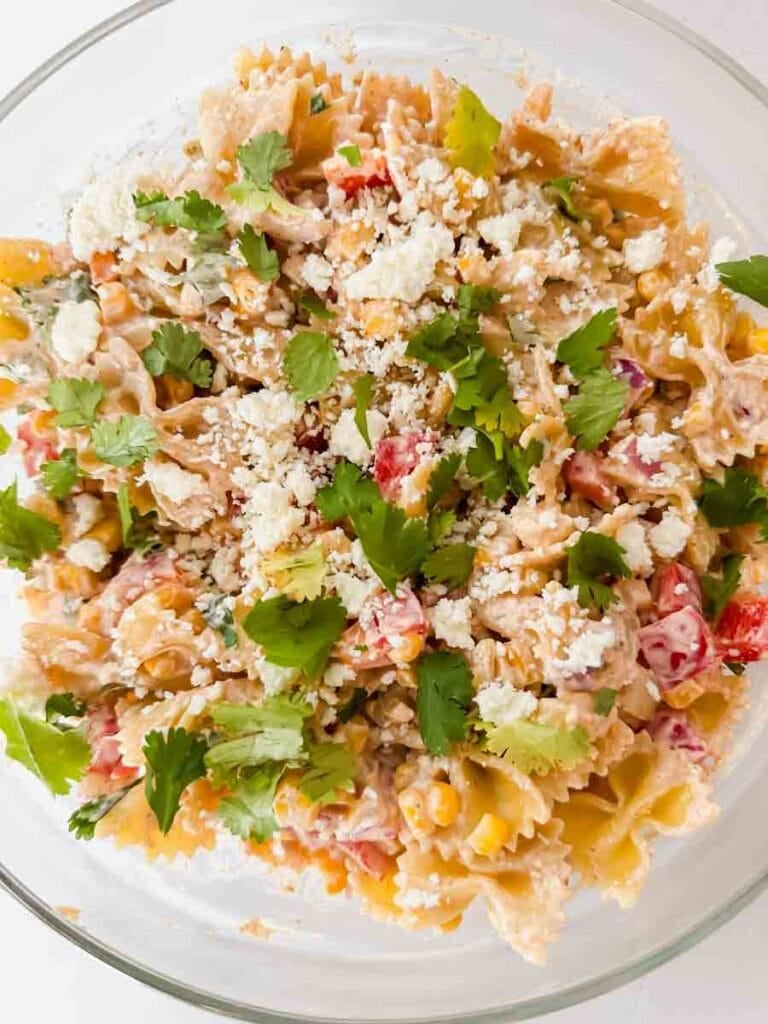 This is a picture of street corn pasta salad. There's a large glass bowl filled with pasta, peppers, corn, cheese, sauce and topped with cilantro and avocado.