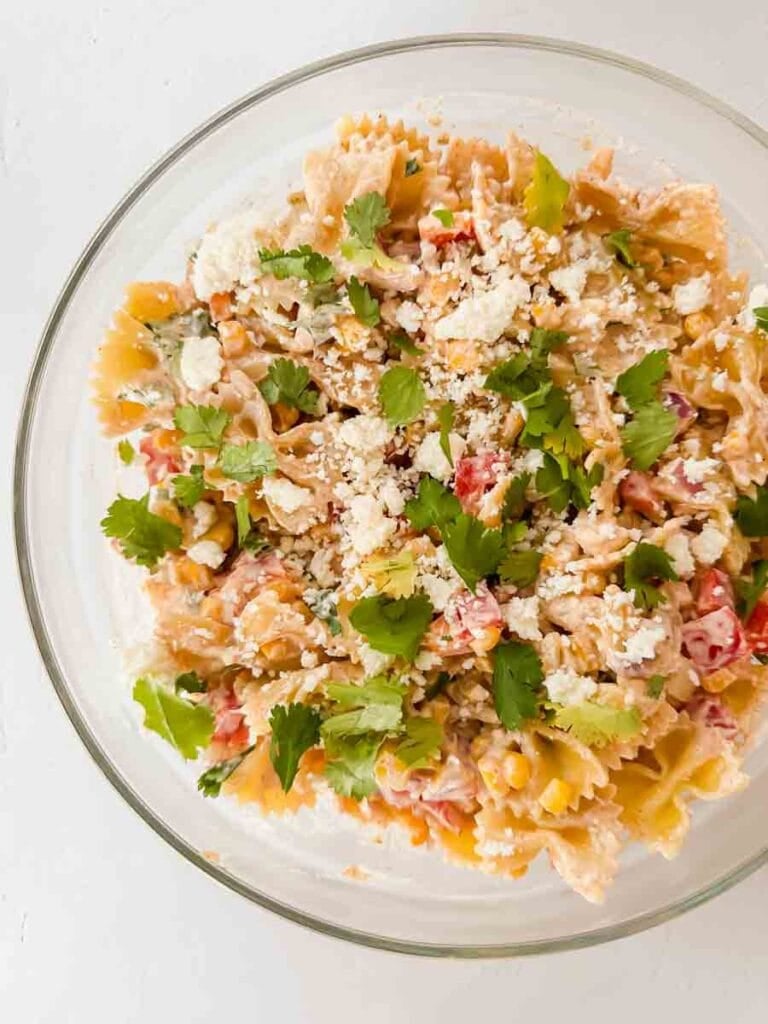 This is a picture of street corn pasta salad. There's a large glass bowl filled with pasta, peppers, corn, cheese, sauce and topped with cilantro and avocado.