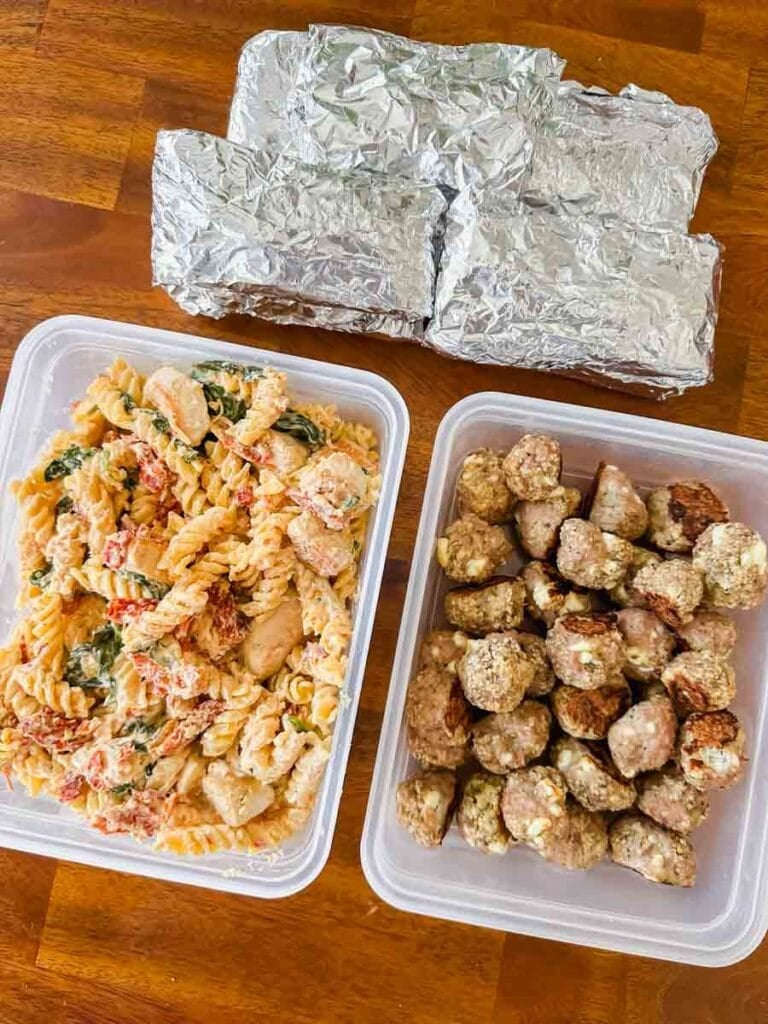 This is a picture of weekly meal prep 48. There's a pile of bacon breakfast burritos stacked together, a big container of marry me chicken pasta, and a big container of greek feta meatballs.