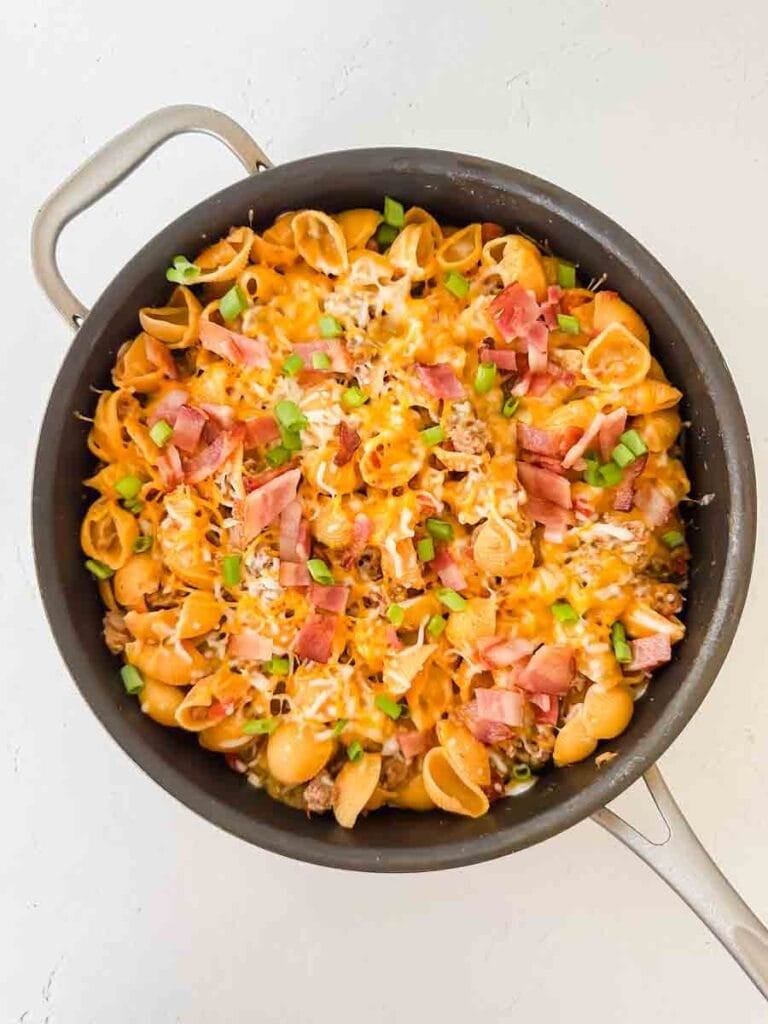 Cheesy pasta with bacon, green onions, and cheese in a large black skillet. This is bacon cheeseburger skillet.