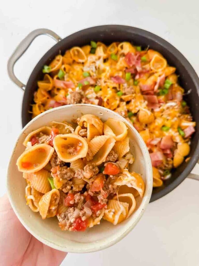 Cheesy pasta with bacon, green onions, and cheese in a large black skillet. This is bacon cheeseburger skillet. A hand is holding up a small ceramic bowl with a portion of the pasta.