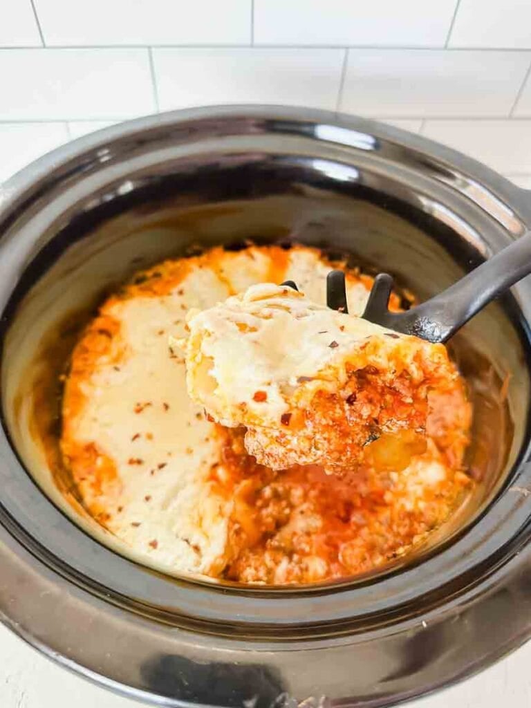 This is a picture of crockpot lasagna. There's a black ceramic insert filled with cheesy lasagna noodles covered in meat sauce. A spoon is holding up a portion.