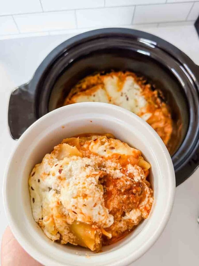 This is a picture of crockpot lasagna. There's a black ceramic insert filled with cheesy lasagna noodles covered in meat sauce. There's a circle white bowl holding up a serving of it.