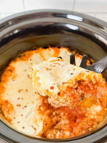This is a picture of crockpot lasagna. There's a black ceramic insert filled with cheesy lasagna noodles covered in meat sauce. A spoon is holding up a portion.