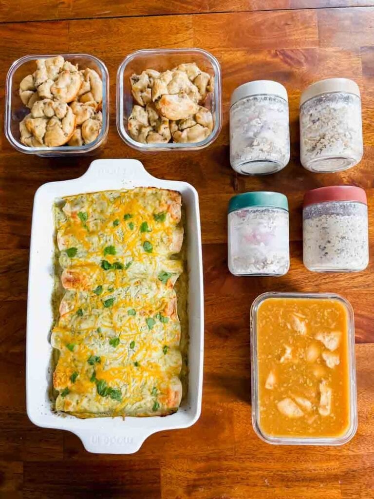 This is a picture of weekly meal prep 49. There's containers of cooked food organized on the table. There's containers of pancake sausage bites, jars of overnight oats, and a big casserole dish of healthy chicken enchiladas.