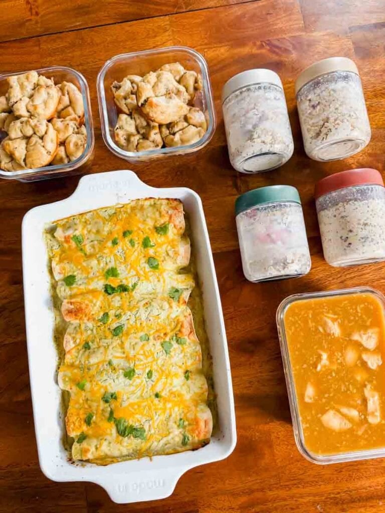 This is a picture of weekly meal prep 49. There's containers of cooked food organized on the table. There's containers of pancake sausage bites, jars of overnight oats, and a big casserole dish of healthy chicken enchiladas.