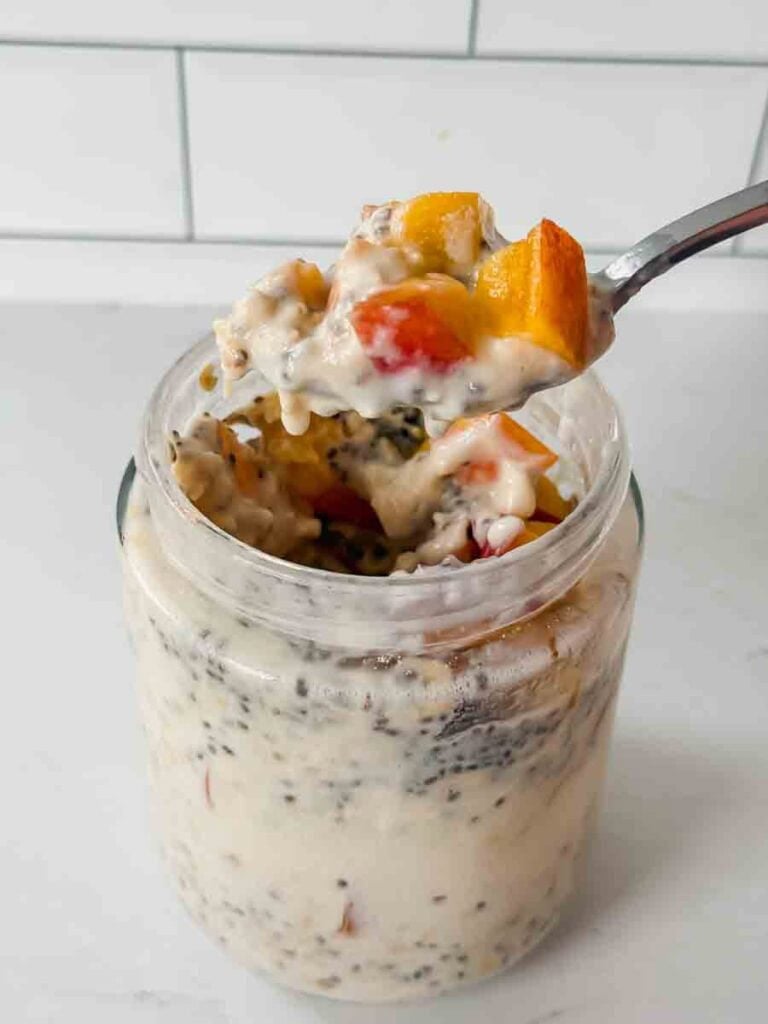 This is a picture of peach overnight oats. There is a glass jar filled with oats, chia seeds, and chopped peaches. A spoon is holding up a bite of the oats.