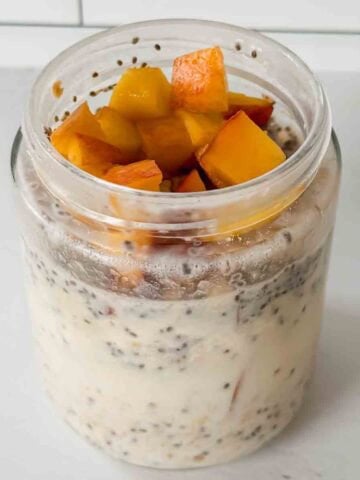This is a picture of peach overnight oats. There is a glass jar filled with oats, chia seeds, and chopped peaches.