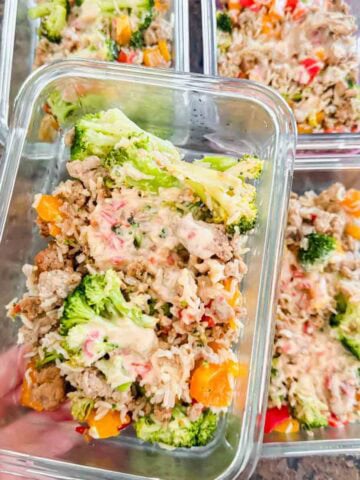 This is meal prep containers of bang bang turkey rice bowls. Each container has ground turkey, rice, broccoli, bell peppers, and a yummy sauce on top. A hand is holding up a container of the bang bang turkey bowl.