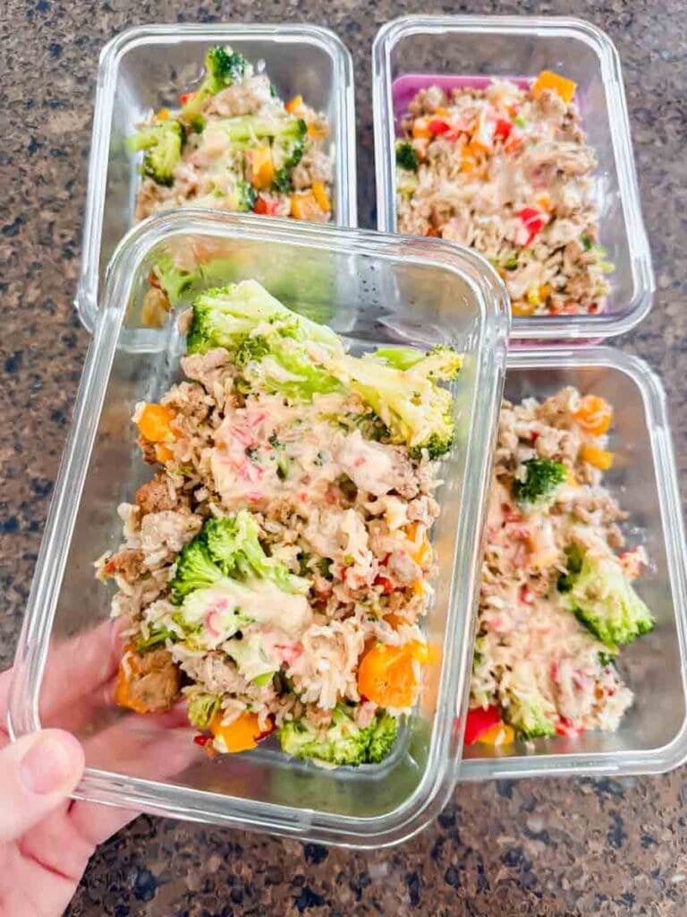 This is meal prep containers of bang bang turkey rice bowls. Each container has ground turkey, rice, broccoli, bell peppers, and a yummy sauce on top. A hand is holding up a container of the bang bang turkey bowl.