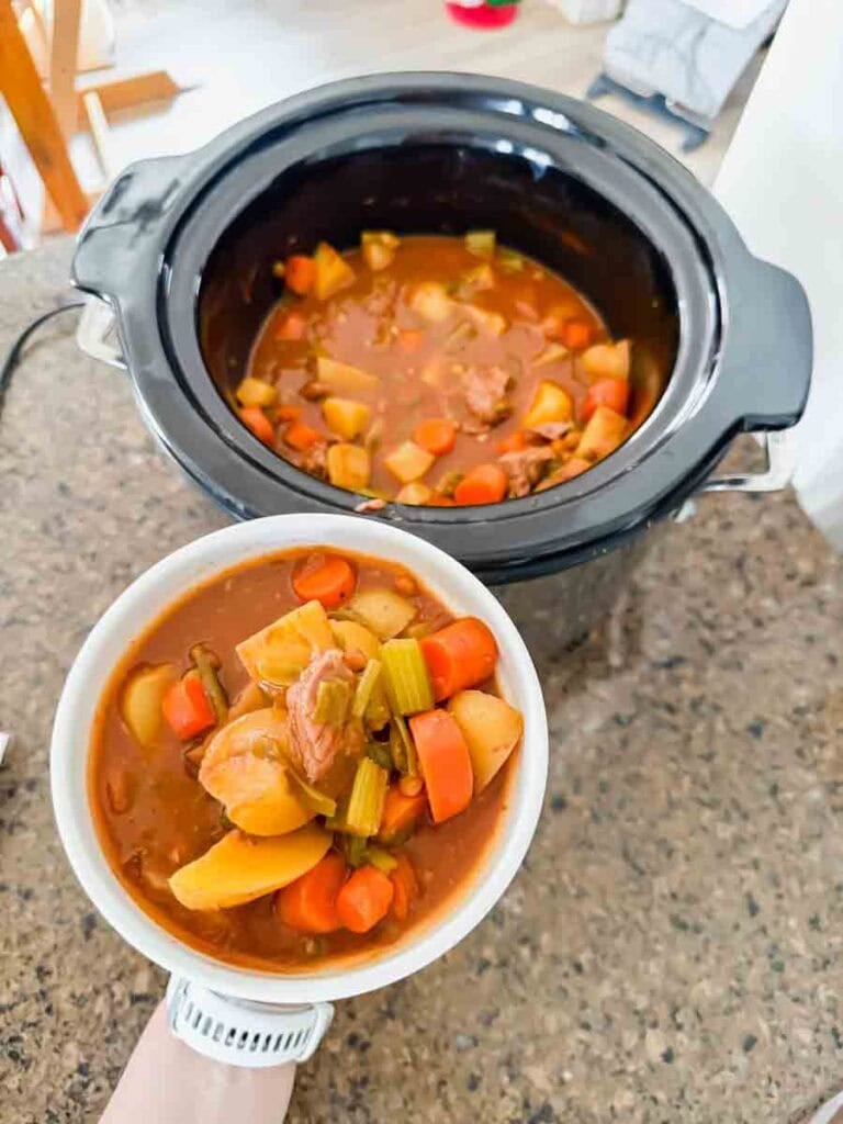 This is a crock pot filled with beef stew. There's beef chunks, potatoes, carrots, green beans, onions, and lentils. The stew is being held by a hand in a white bowl.