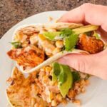 This is a picture of street corn chicken tacos. There's a plate with tortillas, grilled chicken, street corn, and avocado. A hand is holding up the taco as well!