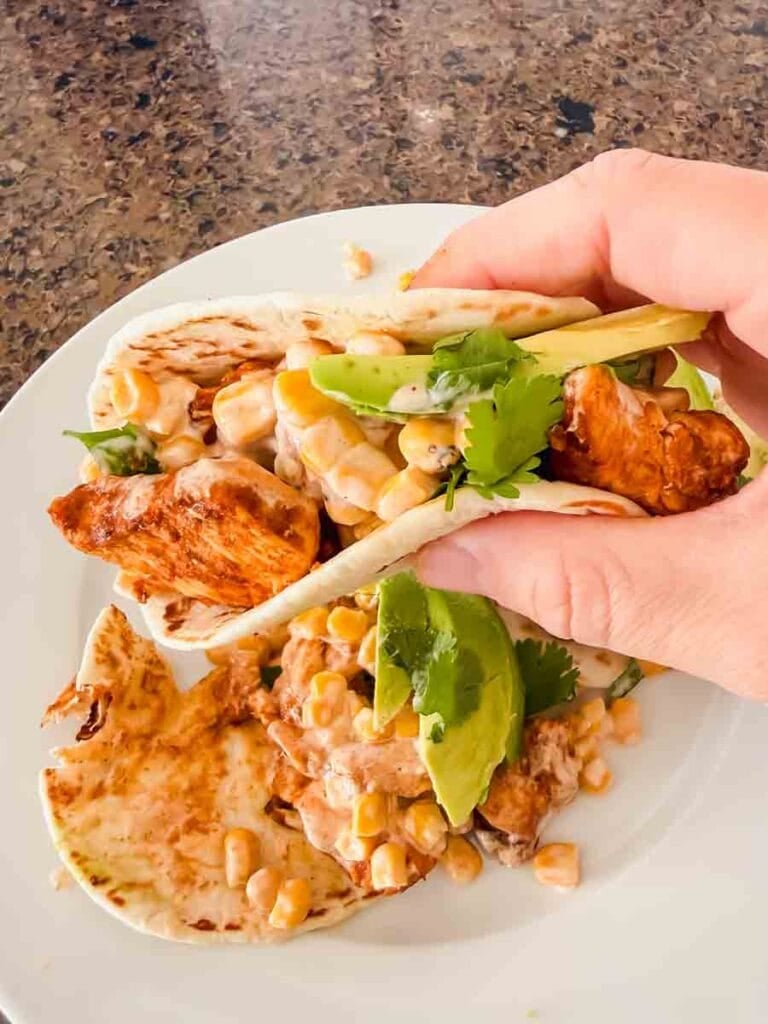 This is a picture of street corn chicken tacos. There's a plate with tortillas, grilled chicken, street corn, and avocado. A hand is holding up the taco as well!