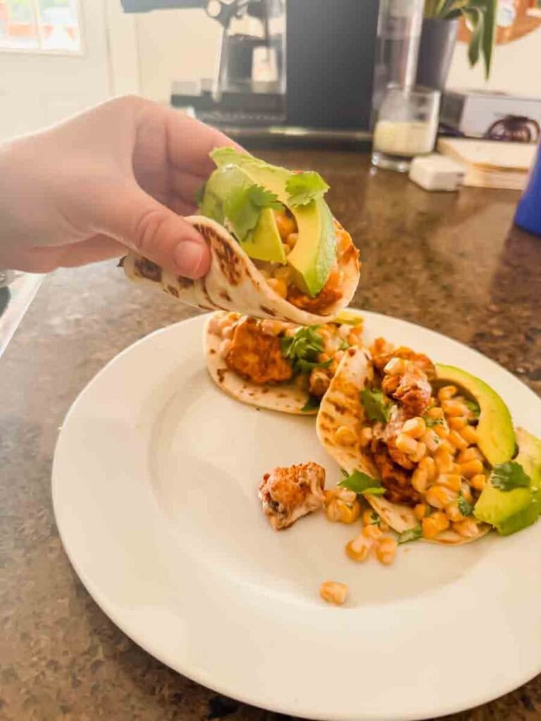 This is a picture of a street corn chicken taco. There's a hand holding up a taco with grilled chicken, street corn, and avocado on top. Another taco is on the plate behind it.
