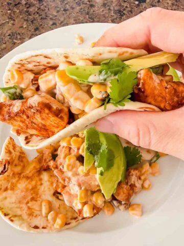 This is a picture of street corn chicken tacos. There's a plate with tortillas, grilled chicken, street corn, and avocado. A hand is holding up the taco as well!