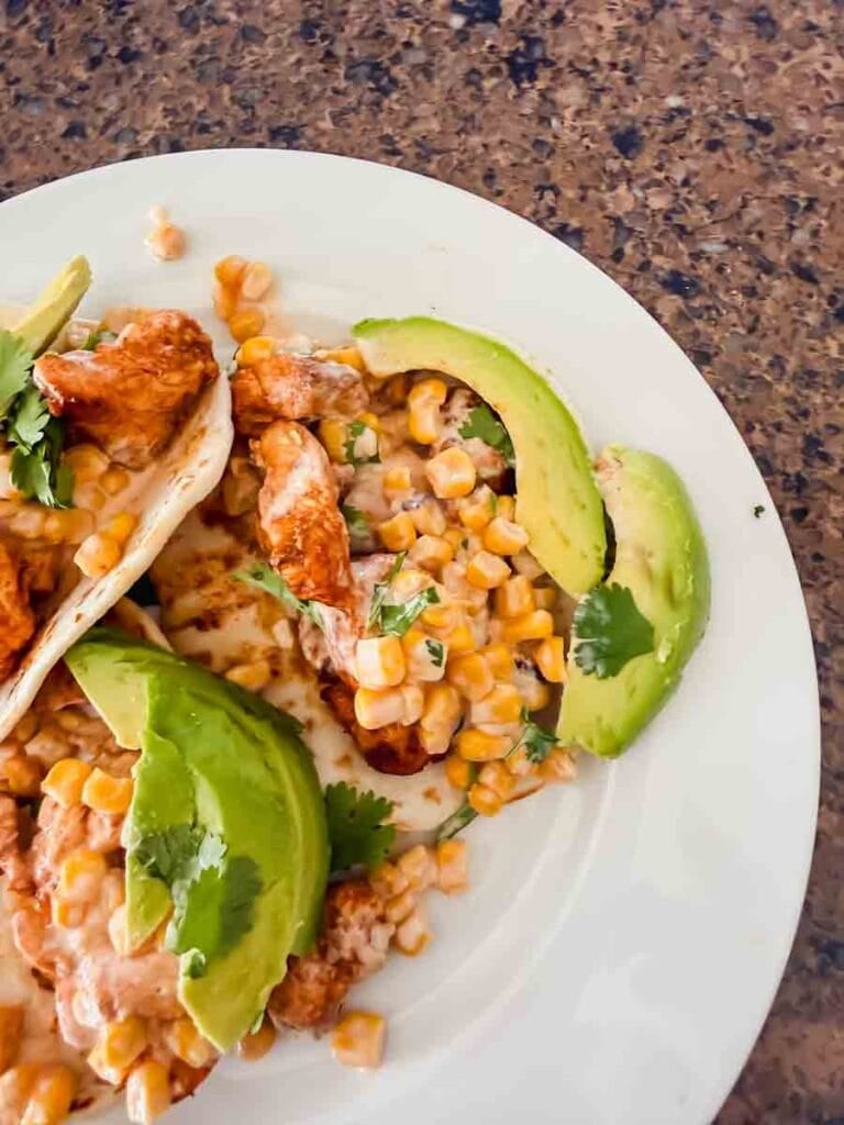 This is a picture of street corn chicken tacos. There's a plate with tortillas, grilled chicken, street corn, and avocado.