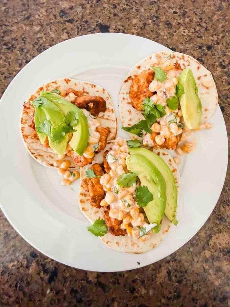 This is a picture of street corn chicken tacos. There's a plate with tortillas, grilled chicken, street corn, and avocado.