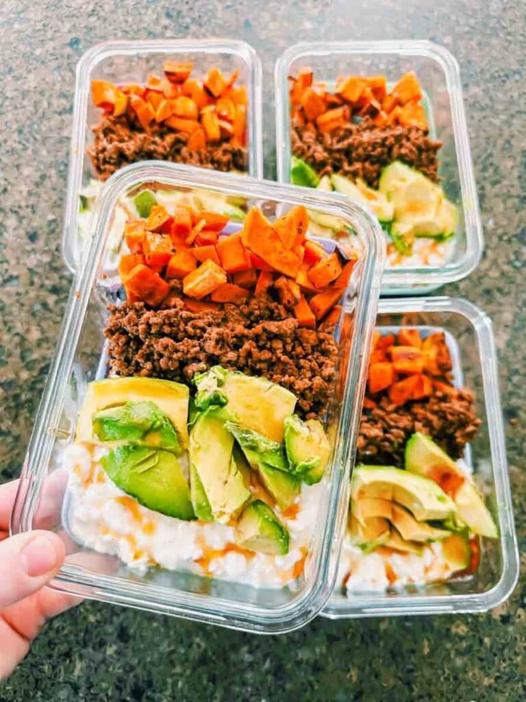 This is a picture of meal prep sweet potato ground beef bowls. There are rectangle glass meal prep containers that have layers of chopped sweet potatoes, ground beef, chopped avocado, and cottage cheese with hot honey drizzled on top. A hand is holding up a container.
