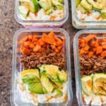 This is a picture of meal prep sweet potato ground beef bowls. There are rectangle glass meal prep containers that have layers of chopped sweet potatoes, ground beef, chopped avocado, and cottage cheese with hot honey drizzled on top.