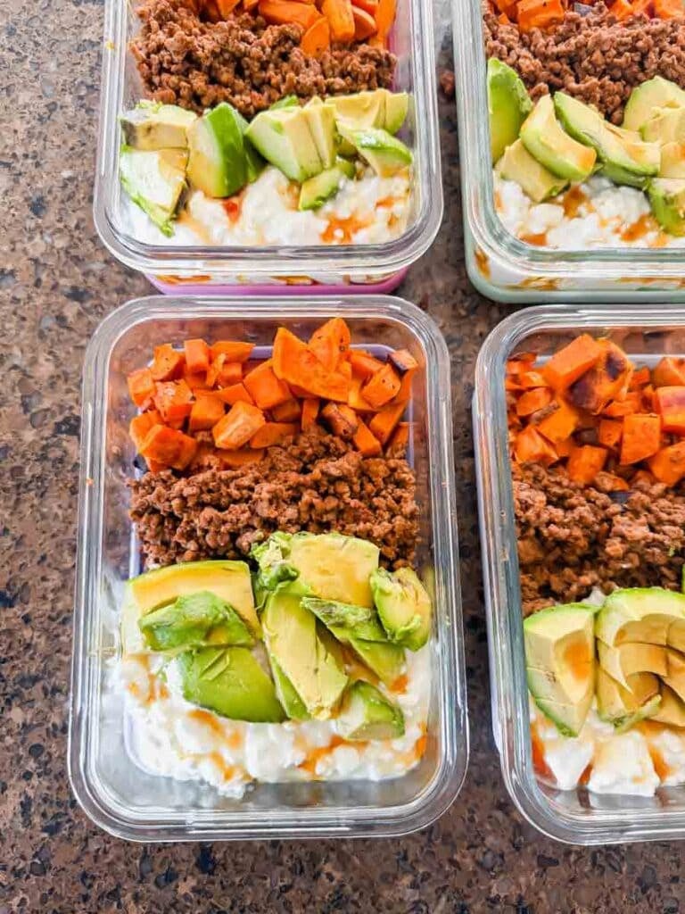 This is a picture of meal prep sweet potato ground beef bowls. There are rectangle glass meal prep containers that have layers of chopped sweet potatoes, ground beef, chopped avocado, and cottage cheese with hot honey drizzled on top.