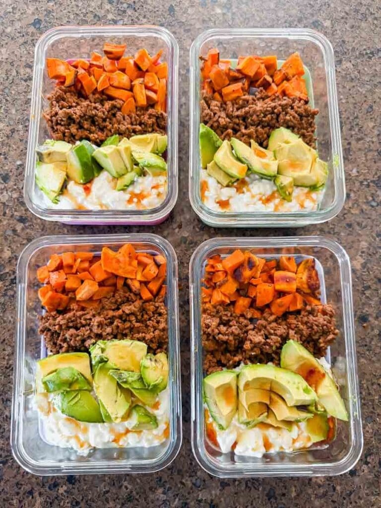 This is a picture of meal prep sweet potato ground beef bowls. There are rectangle glass meal prep containers that have layers of chopped sweet potatoes, ground beef, chopped avocado, and cottage cheese with hot honey drizzled on top.